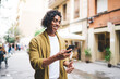 © BullRun - Smiling curly handsome hipster guy reading message on mobile phone enjoying free time on city street, positive young male influencer share content in blog strolling during journey on urban settings