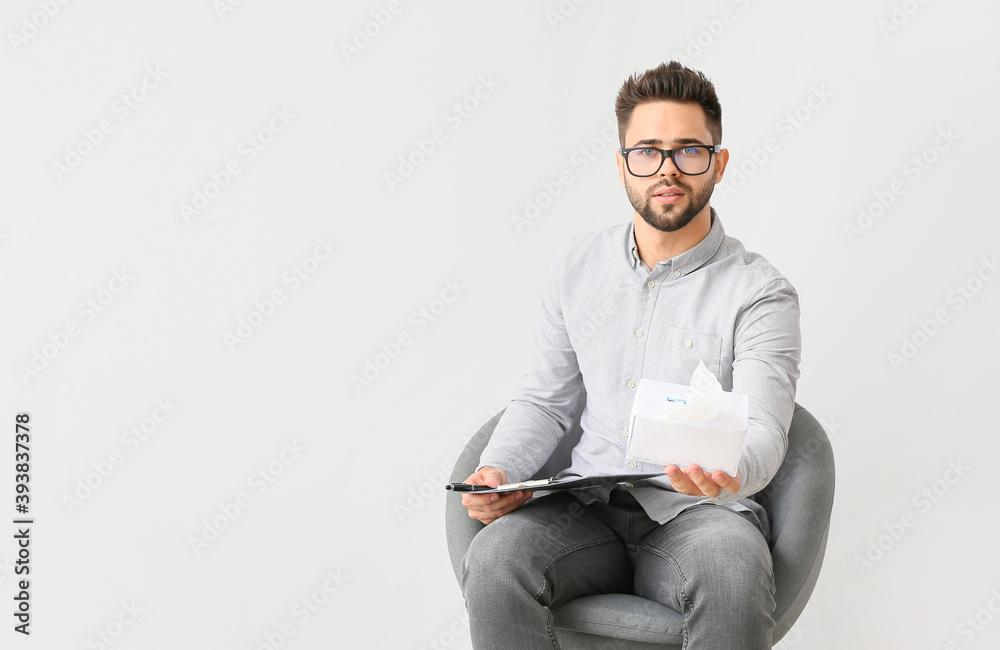Male psychologist with tissues sitting in armchair on light background