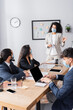 © LIGHTFIELD STUDIOS - Hispanic businesswoman in medical mask pointing with finger and looking at co-workers during meeting in office