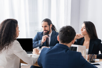 Wall Mural - Hispanic businessman looking at colleague near devices on blurred foreground in office