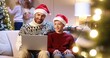 © VAKSMANV - Portrait of happy Caucasian teen boy sitting with dad in santa hat at living cozy room and typing on laptop choosing gifts Mom and small kid decorating Christmas tree on background Holiday preparation