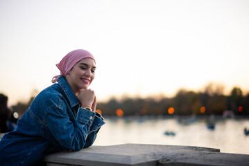 Young woman smiling fighter against cancer with pink scarf on her head observing the tranquility of the lake at sunset