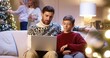 © VAKSMANV - Happy Caucasian dad with teen son sitting in cozy room typing on laptop spending holiday time on internet buying gifts. Mom and small kid decorating glowing Christmas tree. Family holidays concept