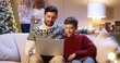 © VAKSMANV - Happy Caucasian dad with teen son sitting in cozy room typing on laptop spending holiday time on internet buying gifts. Mom and small kid decorating glowing Christmas tree. Family holidays concept