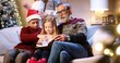 © VAKSMANV - Happy Caucasian family at home on Christmas Eve. Cute kids sitting on sofa with grandpa and reading book while parents decoration xmas glowing tree on background. Happy holidays concept