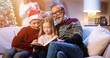 © VAKSMANV - Portrait of joyful Caucasian cute children talking and reading book with grandfather sitting at home on Christmas Eve while parents decoration xmas glowing tree on background New Year concept