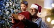 © VAKSMANV - Close up portrait of joyful Caucasian old grandpa in santa hat with grandchild sitting in decorated room typing and browsing on tablet while talking near glowing new year tree. Xmas holidays concept