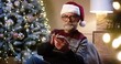 © VAKSMANV - Close up portrait of old grey-haired Caucasian male pensioner grandpa in santa hat typing on smartphone buying gifts online while resting in cozy room near glowing christmas tree. Holidays concept