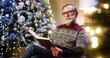 © VAKSMANV - Portrait of old nice Caucasian male pensioner grandpa in glasses and christmas sweater reading book while sitting in decorated room near glowing christmas tree. Xmas magic. Winter holidays concept.