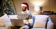 © VAKSMANV - Portrait of happy Caucasian handsome male in santa hat sitting in cozy room with xmas tree and typing on laptop looking away. Happy holidays. Winter concept. New Year's Eve. E-commerce