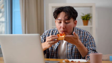 Young asian man biting slice of pizza when reading e-mail from colleague on laptop screen