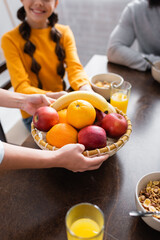 Wall Mural - Cropped view of woman holding basket with fresh fruits near smiling daughter and husband in kitchen