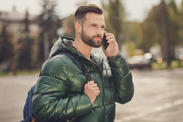 Poster - Photo of cheerful cute young guy dressed green coat rucksack walking talking modern gadget outdoors city street