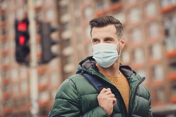 Poster - Photo of handsome good mood young guy dressed green coat rucksack protection mask walking outdoors urban city street