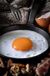 Close-up of frame-filling pan containing a fried egg, with mushrooms and bacon in the foreground, cloth, fork and egg-shell behind, with directional light from the side.