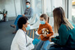 © Drazen - Black female pediatrician talking to mother and daughter in a waiting room during coronavirus pandemic.