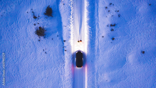 Aerial top view of romantic couple of tourists standing near car while headlights lighting them way on in winter darkness, bird\'s eye view of vehicle in snowy north lands. Wanderlust in road trip
