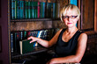 © pavasaris - Close-up portrait of a middle-aged businesswoman with glasses next to bookshelves, smiling