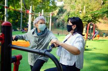 A female physiotherapist or professional caregiver helps a senior adult woman to get on an air swinger outdoor machine. Both wearing face medical masks because of the coronavirus pandemic.