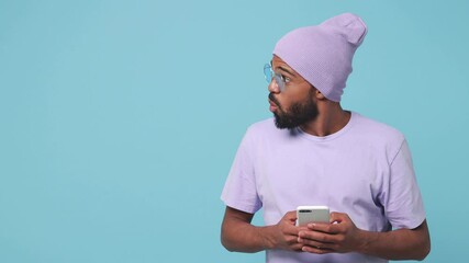 Wall Mural - Shocked young african american man in violet t-shirt hat isolated on blue background studio. People lifestyle concept. Using mobile cell phone typing sms message looking aside say wow showing thumb up