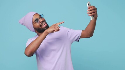 Wall Mural - Funny african american man in t-shirt hat isolated on blue background studio. People lifestyle concept. Doing selfie shot on mobile phone making video call showing biceps victory sign point on himself