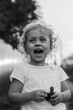 A little blond curly girl is standing near the fence in the countryside in summer with laugh and open mouth black and white, vertical