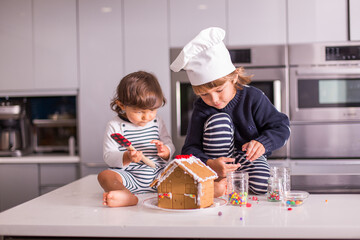 Cute children sibling boys preschooler and  toddler enjoying making a Christmas gingerbread house in the kitchen during a pandemic holiday season baking and decorating using colorful candy.