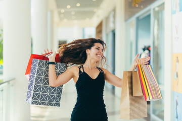 Cheerful lady in stylish black dress carrying on colorful shopping bags while walking at store. Attractive brunette looks satisfied after shopping at mall.