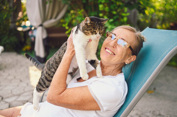 Wall Mural - Happy smiling senior elderly woman in glasses relaxing in summer garden outdoors hugging domestic tabby cat. Retired old people and animals pets concept