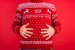 Holidays overeating concept. Close up photo of men in red winter sweater with deers holding his big tummy isolated on red background