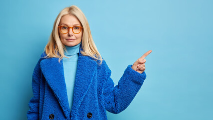 Horizontal shot of serious self confident blonde woman wears spectacles and winter coat points away on blank space demonstrates place for advertisement or promo attracts your attention to item