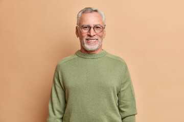 Wall Mural - Half length shot of cheerful senior man smiles happily with white teeth wears optical glasses and sweater isolated over brown background. Happy grandfather glad grandchildren came to visit him