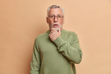 Wall Mural - Surprised bearded grey haired speechless man holds chin and stares bugged eyes hears secret or shocking news dressed in basic long sleeved jumper isolated over brown background holds breath from awe