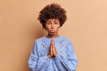 Curly haired sad woman purses lips keeps palms pressed together and looks unhappily imploring at camera asks for apology needs help poses over brown background feels guilty for doing something wrong