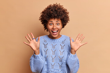Wall Mural - Overjoyed dark skinned woman has curly Afro hair feels very happy raises palms reacts on awesome surprise wears knitted sweater poses against brown studio background. Wow what amazing thing!