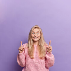 Wall Mural - Happy glad European woman with blonde hair smiles pleasantly indicates above on blank space gives place for your advertisement wears knitted sweater isolated over purple background. Check this out
