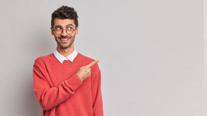 Studio shot of handsome cheerful man with bristle smiles pleasantly points sideways on blank space shows advertisement smiles gladfully wears spectacles neat red sweater isolated over grey wall