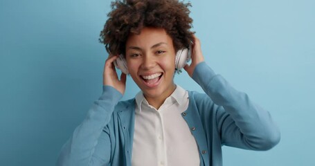 Wall Mural - Joyful optimistic Afro American woman listens pleasant music in stereo headphones closes eyes and laughs moves with rhythm of song poses against blue background. People lifestyle hobby concept