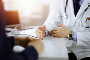 A doctor is talking to his patient, while sitting together at the desk in the sunny cabinet in a hospital. Physician using clipboard for filling up medication history records. Perfect medical service