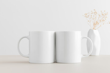 Sticker - Two white mugs mockup with a gypsophila in a vase on a beige table.