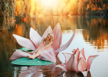 Happy Fantasy Young Blonde Woman Little Fairy Princess Sitting In Pink Lotus Flower On Lake Water. Elf Girl, With Smiling Face. Autumn Nature Background Orange Trees Divine Magic Sun Light. Pink Dress