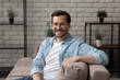 © fizkes - Portrait of smiling young 30s handsome man in eyeglasses relaxing on comfortable sofa in modern living room, happy guy enjoying free weekend leisure time chilling alone at home, looking at camera.