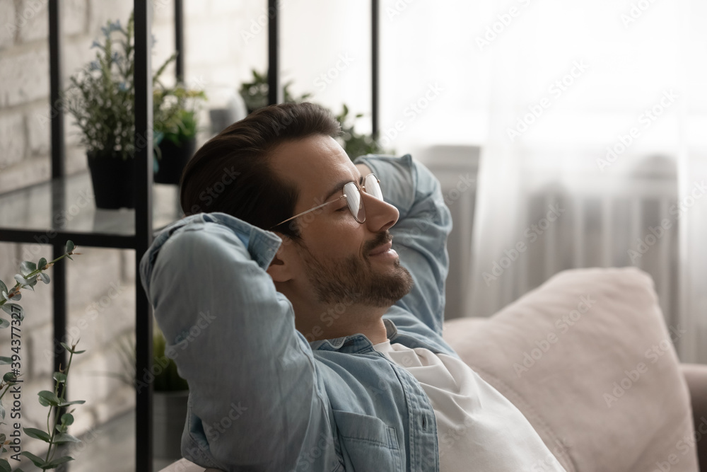 Side head shot view calm serene young 30s man in eyeglasses resting on cozy sofa with closed eyes and folded behind head hands, daydreaming sleeping alone or visualizing positive future at home. - obrazy, fototapety, plakaty 