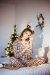 Happy young adult woman wake up at morning in cozy bedroom with decorated christmas tree, raised hands up, making smiling face