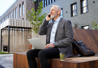 Creating strong relationships. Busy elegant mature man talking on the phone, working on his laptop while sitting on the bench outdoors