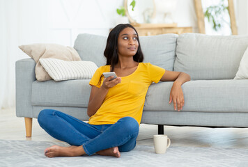 Wall Mural - Remote education, online work concept. Thoughtful black woman with cellphone sitting on floor at home, copy space