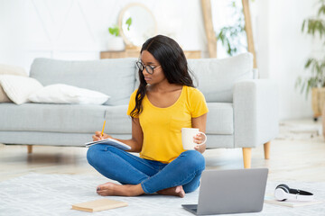 Wall Mural - Smart black female student taking notes during online lecture on laptop, studying on floor at home, full length portrait