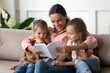 © fizkes - Caring young mother reading interesting book to little preschool daughters, family sitting on comfy couch in living room enjoy fairytale story spend time together. Development of kids activity concept