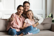 © fizkes - Loving young man hug his beloved wife and little daughter sit on couch in living room, happy people smiling looking at camera posing for photo picture. Exemplary family portrait, love and bond concept