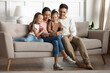 Married couple with 2 preschool adorable daughters resting seated on couch in living room with modern gadget having fun using smartphone. Take family picture, make videocall, parental control concept